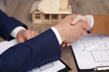 Real estate agent and client shaking hands at desk with house model in office, closeup