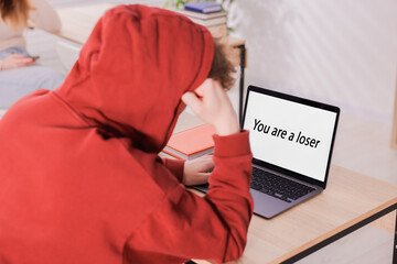 Cyber bullying. Upset teenage boy and laptop with text You Loser at desk indoors, closeup