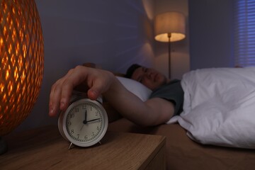 Frustrated man suffering from insomnia and turning off alarm clock in bed indoors, selective focus