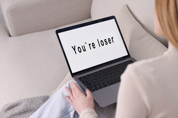 Cyber bullying. Upset teenage girl looking at laptop with text You Loser on sofa at home, closeup