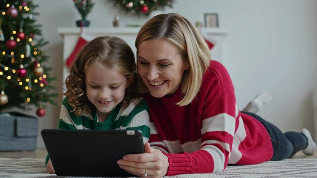 Smiling mother and daughter lie on the rug near a Christmas tree, looking at a tablet. They are wearing cozy sweaters. The holiday season provides a setting for family bonding