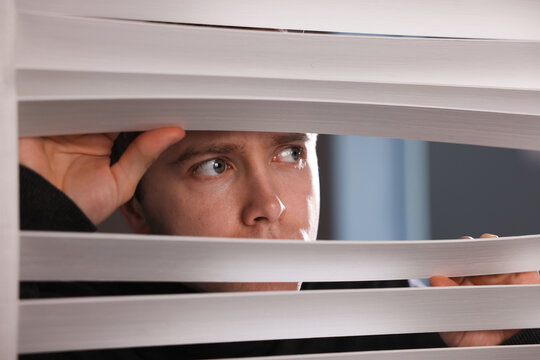 Paranoia. Worried man looking through window blinds indoors