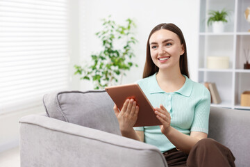 Young woman using modern tablet in armchair at home. Space for text