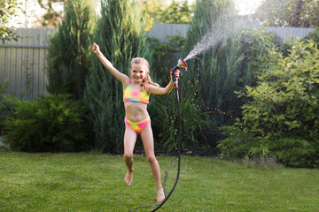 Happy little  girl playing with garden hose and having fun with spray of water in sunny backyard....