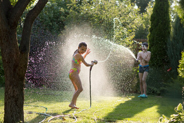 Happy children brother and sister playing in the garden hose and having fun with spray of water in...