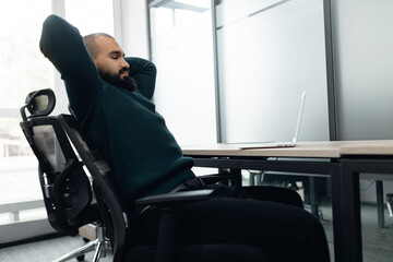 Professional man with beard sitting comfortably in modern office, arms behind his head, taking...