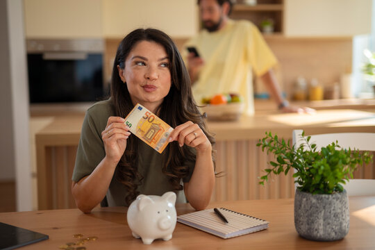 Smiling woman holding a 50 euro banknote next to a piggy bank, sitting at a kitchen table with a notebook and coins. A man in the background looks at his phone, suggesting a casual home scene focused 