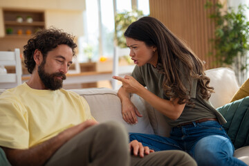 Angry young woman pointing her finger at a man during a heated argument at home. The man looks down, showing discomfort or guilt, while sitting on a sofa in a modern living room.
