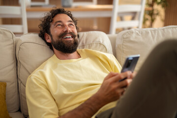 Smiling man lying on a sofa and holding a smartphone, enjoying a lighthearted moment at home. Relaxed and cozy indoor scene with a casual atmosphere.
