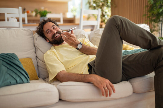 Cheerful man lying on a sofa and using speakerphone on his smartphone. Relaxed atmosphere in a cozy home, enjoying a casual conversation and leisure time.
