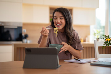 Cheerful young woman eating noodles while watching a tablet at the kitchen table, with a notebook...