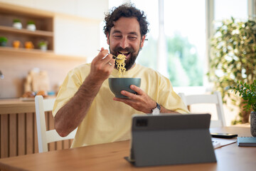 Smiling man eating noodles from a bowl while sitting at the kitchen table with a tablet in front of him.
