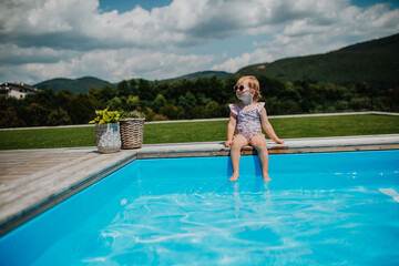 Little girl sitting by pool, splashing with legs in water.