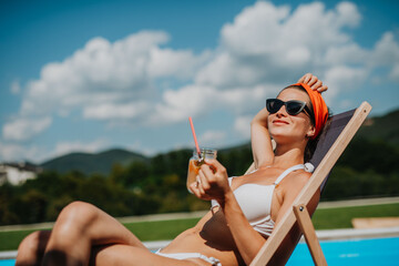 Young woman sitting by swimming pool and drinking limonade.