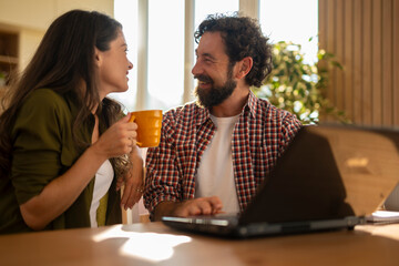 Happy couple sitting at the table with a laptop, smiling and looking at each other while the woman holds a mug.
