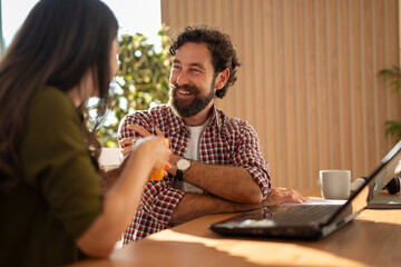 Smiling man sitting at a table with a laptop, enjoying a conversation with a woman holding a mug at home.
