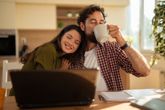 Loving couple sitting together at home, woman resting her head on man's shoulder while he drinks coffee and works on a laptop.