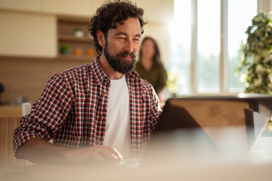 Young man working on a laptop at home, focused and smiling, with a woman blurred in the background.
