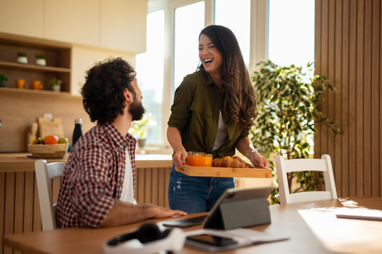 Happy young couple spending time together in a bright modern kitchen. Woman is serving breakfast while the man works on a tablet. Casual home setting with natural light and relaxed atmosphere. Remote
