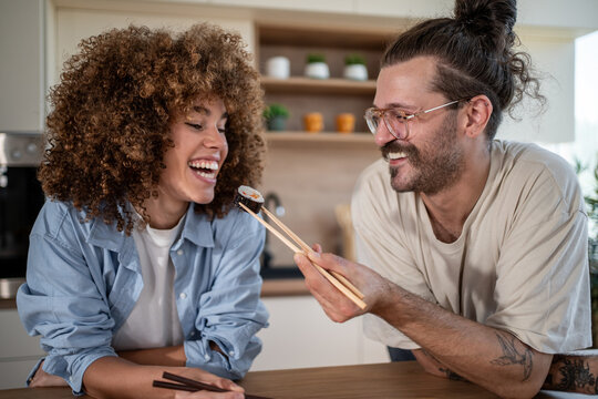 Happy couple sharing sushi in modern kitchen: feeding each other and smiling