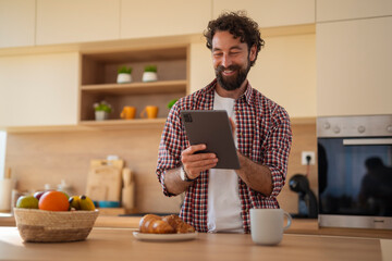 Bearded man in a casual shirt smiling while using a tablet in a cozy kitchen setting.
