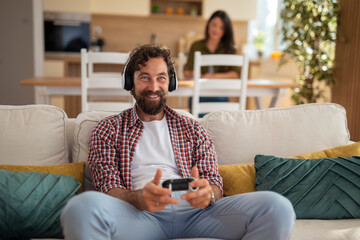 Obraz premium Happy bearded man playing video games at home while his partner watches from the background. 