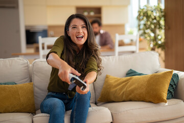 Happy young woman playing video games enthusiastically while sitting on couch at home.
