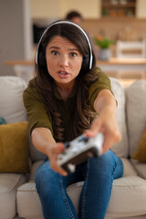 Focused young woman wearing headphones and playing video games with controller at home.
