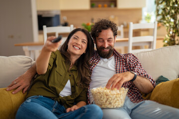 Happy young couple cuddling on couch and watching TV while eating popcorn at home.