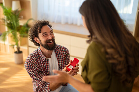 Smiling man proposing to woman with engagement ring box in hand at home.