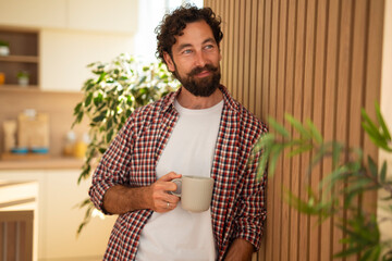 Relaxed young man holding coffee mug and standing by kitchen wall at home.
