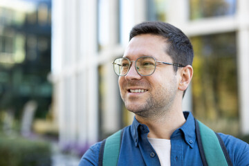 A smiling man wearing glasses and a backpack poses outdoors in front of a building on a sunny day.