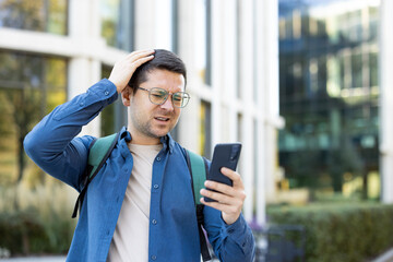 A man is looking at his phone with a confused expression, touching his head, near a modern building...