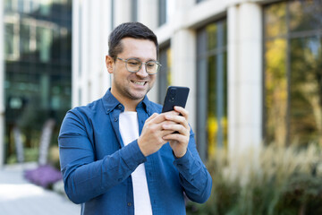 A smiling man in glasses is using his smartphone outdoors in front of a building. The man is happy.