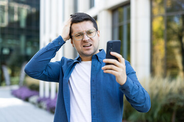 A concerned man holds his head while looking at his phone outdoors near a building.
