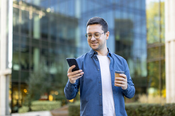 A man in glasses smiles while looking at his phone, holding a coffee cup in front of a modern...