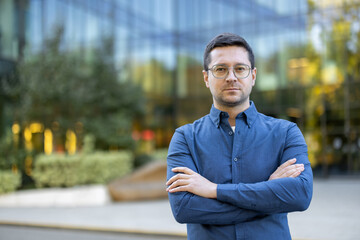 A man in a blue shirt and glasses stands in front of a modern building with his arms crossed,...