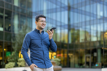 A businessman in glasses talks into his phone while standing outside a modern building.