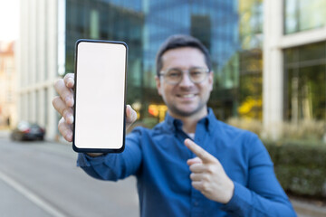 A man holds a phone with a blank screen, pointing at it in a business district.