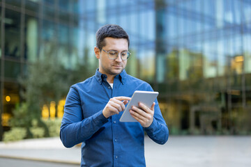 A man in glasses and a blue shirt, focused on a tablet in front of a modern building.