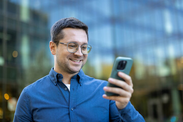 A smiling man in glasses looks at his phone in front of a modern building.