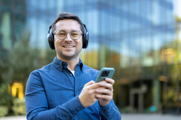 A man wearing headphones and glasses smiles while using his phone outdoors, with a building in the...