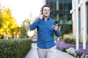 A man in a blue shirt listens to music on headphones while walking outside holding his phone.