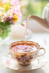 Creative postcard with porcelain cup of tea, teapot and a vibrant bouquet of garden flowers. Elegant tea set in beautiful morning sun light. Good morning and teatime concept. Slow life visual trend.