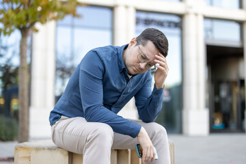 A man in a blue shirt sits outdoors, looking down with a concerned expression, holding his head.