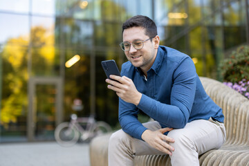 A smiling man in glasses checks his phone while sitting on a bench outdoors near an office building.