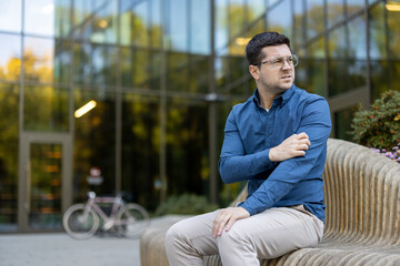 A man sits on a bench outside, scratching his arm near a modern glass building and a bicycle,...