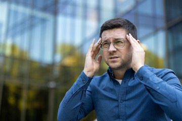 A man with a headache holds his temples, experiencing pain. He is outdoors, near a building,...