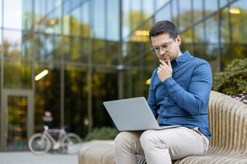 A man with glasses thoughtfully looks at his laptop while seated on a bench outdoors, near a modern...