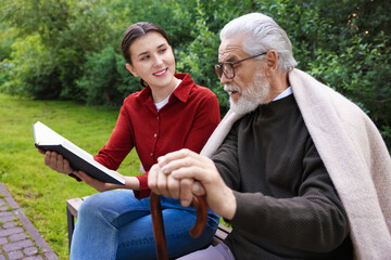 Smiling caregiver and elderly man in glasses with walking cane reading book on bench outdoors